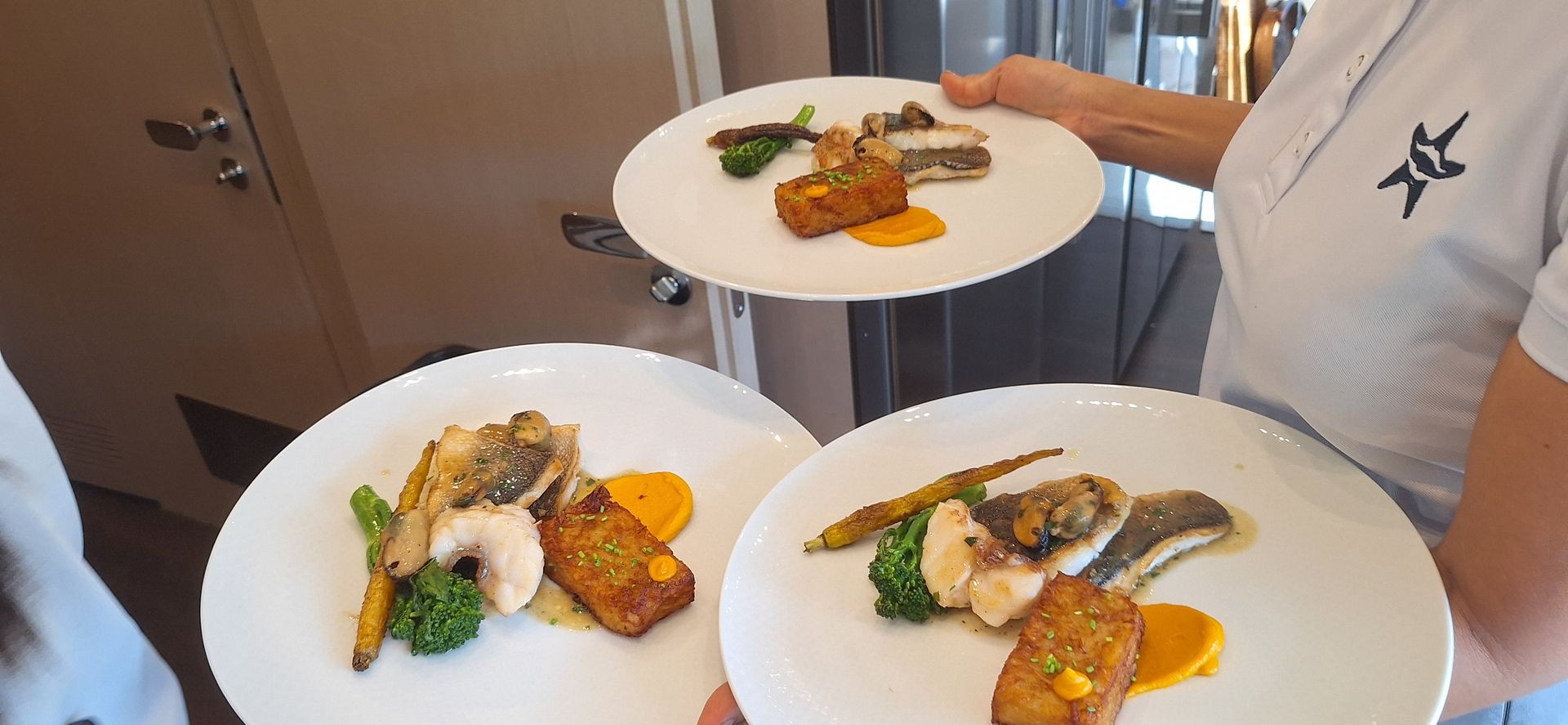 Server holding three plates of gourmet fish with vegetables and sauce.